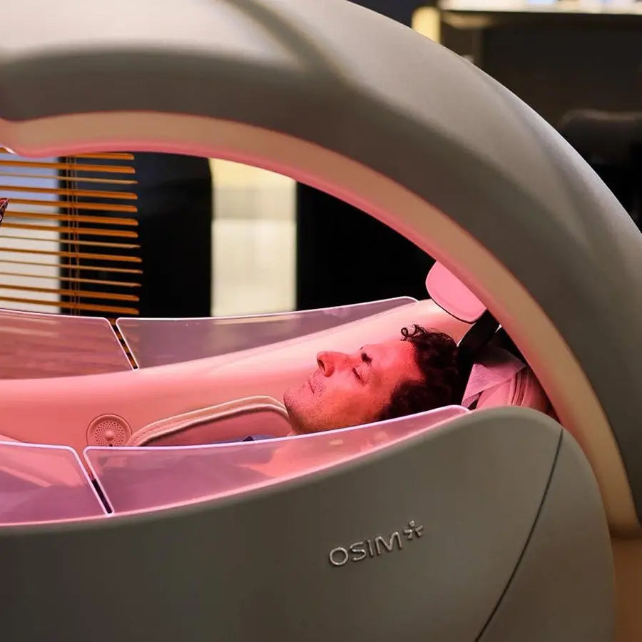 A man reclining inside an premium OSIM uDream·AI massage chair under a curved dome, with pinkish-red light illuminating his face and upper body.