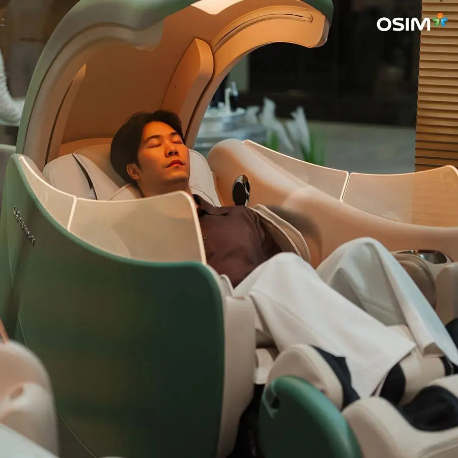 A man reclines peacefully inside a large, futuristic OSIM massage chair in a modern indoor setting. The chair features a curved design with a soft mint green and white color scheme, and the OSIM brand logo is visible in the top right corner.