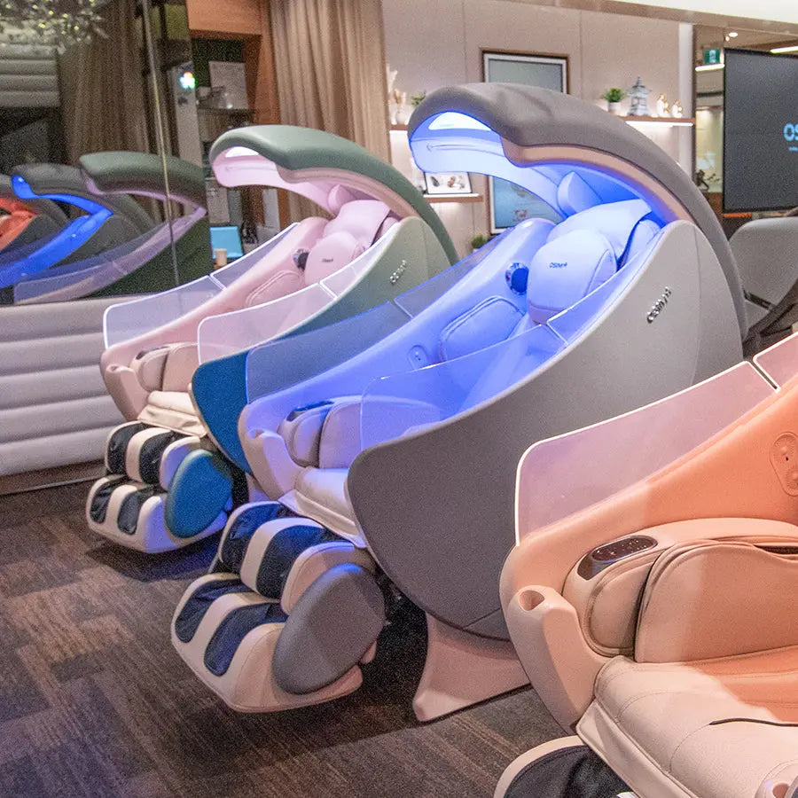 A row of modern pod-style massage chairs in different colors, including gray, peach, blue, and sage green, arranged on a dark wood floor inside a showroom.