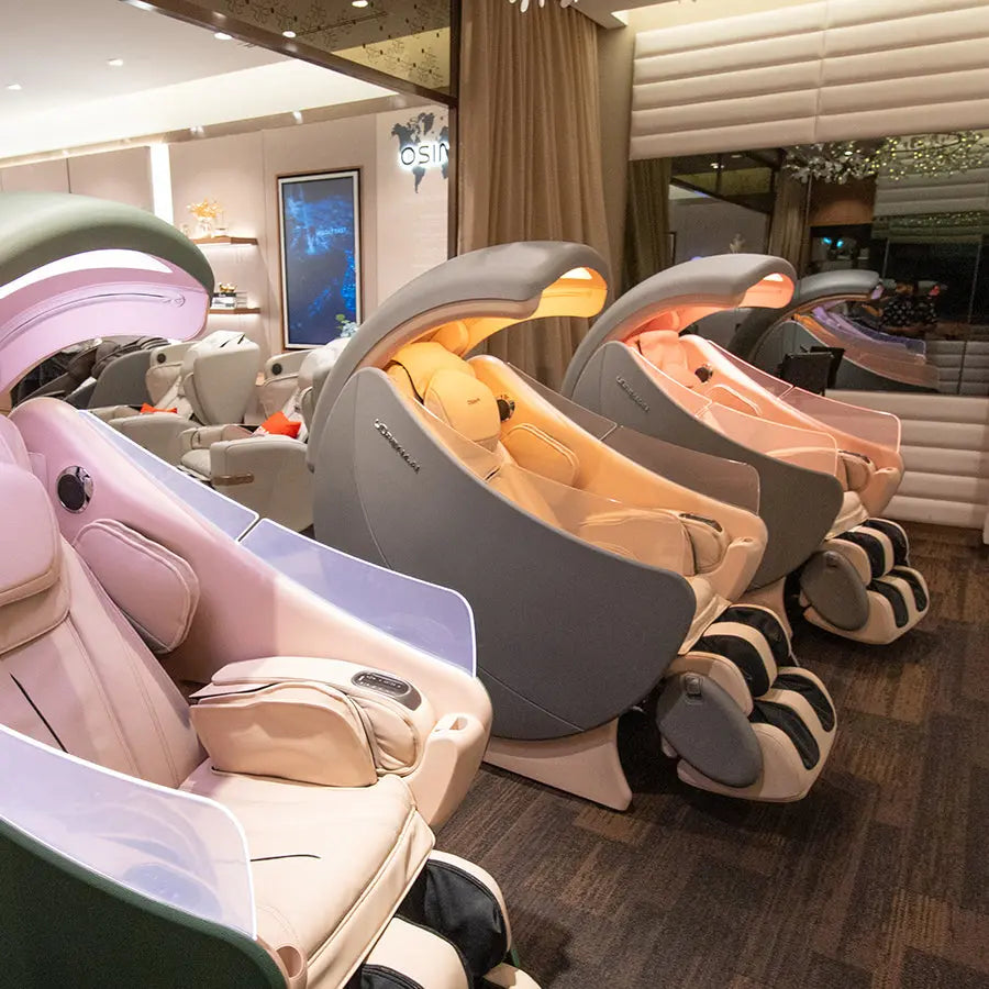 A showroom interior featuring multiple modern massage chairs in soft pastel colors including pink, gray, and beige, arranged along a dark wood floor.